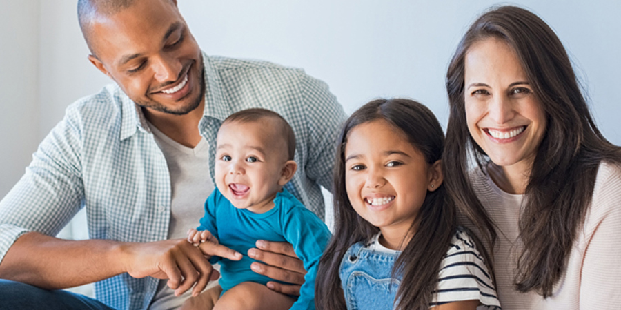 Young family smiles at camera. Dad is holding baby and mom sits with older daughter.